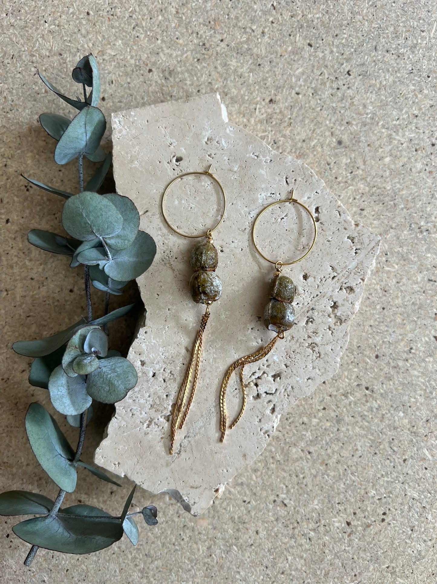 Pair of gold hoop earrings with green beads on a textured stone surface with eucalyptus leaves.