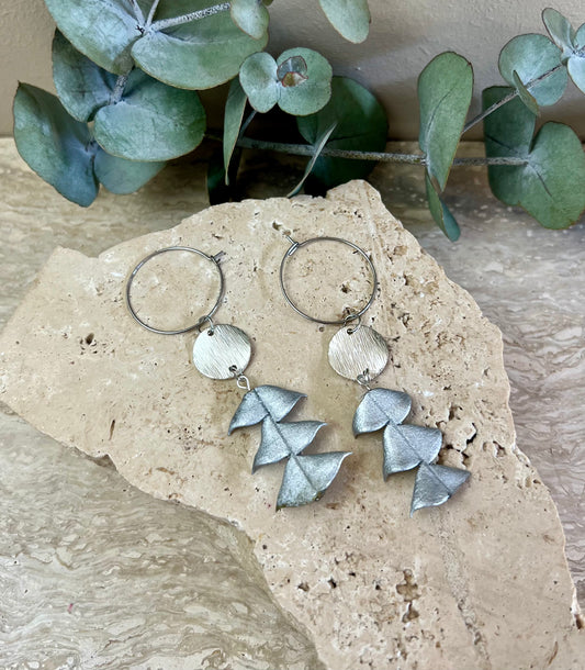 Silver hoop earrings with leaf-shaped pendants on a stone surface with eucalyptus leaves in the background.