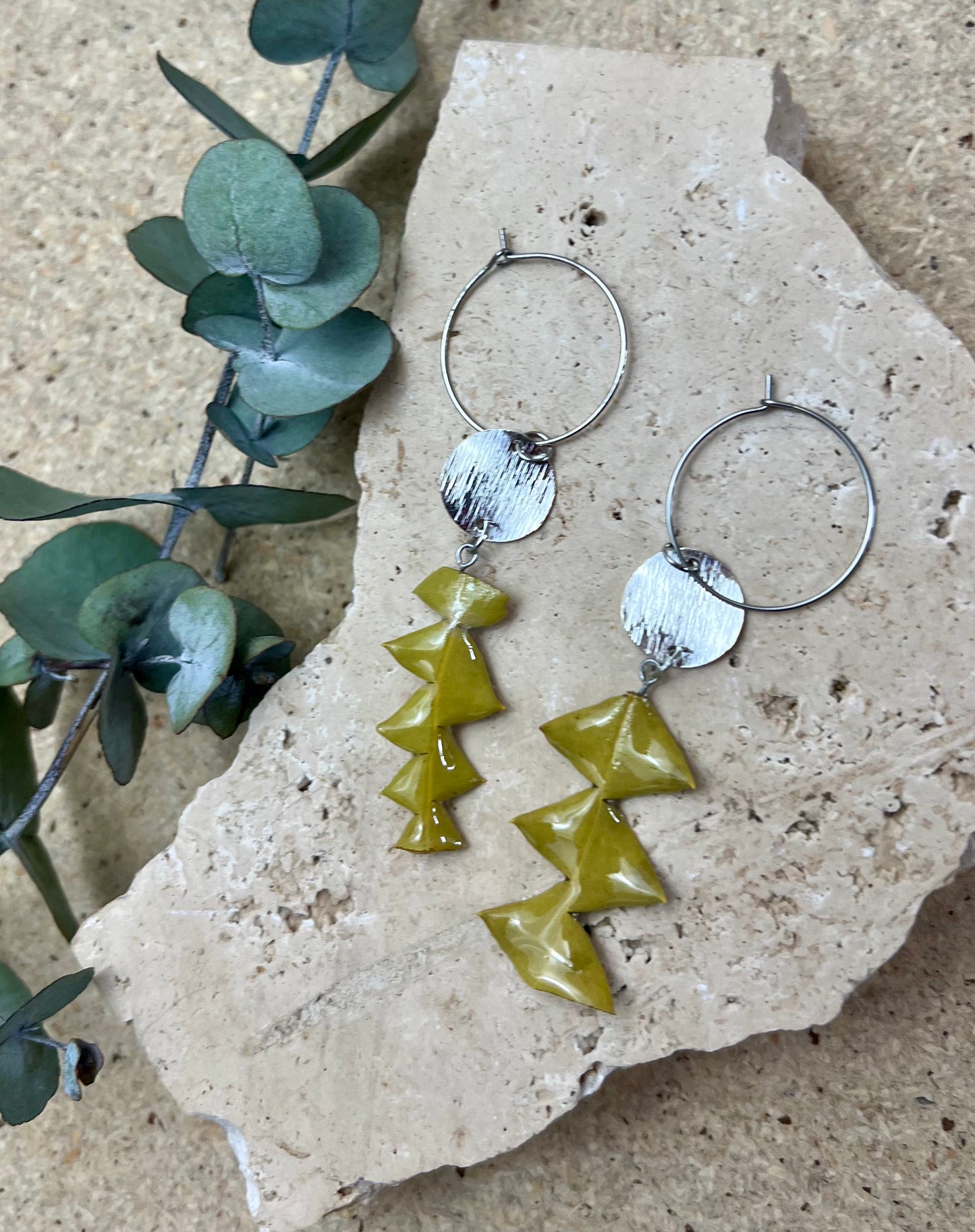 Pair of hoop earrings with green and silver geometric pendants on a stone surface.