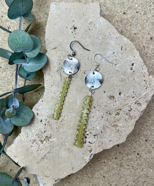 Silver earrings with leaf-like pendants on a stone surface with eucalyptus leaves.