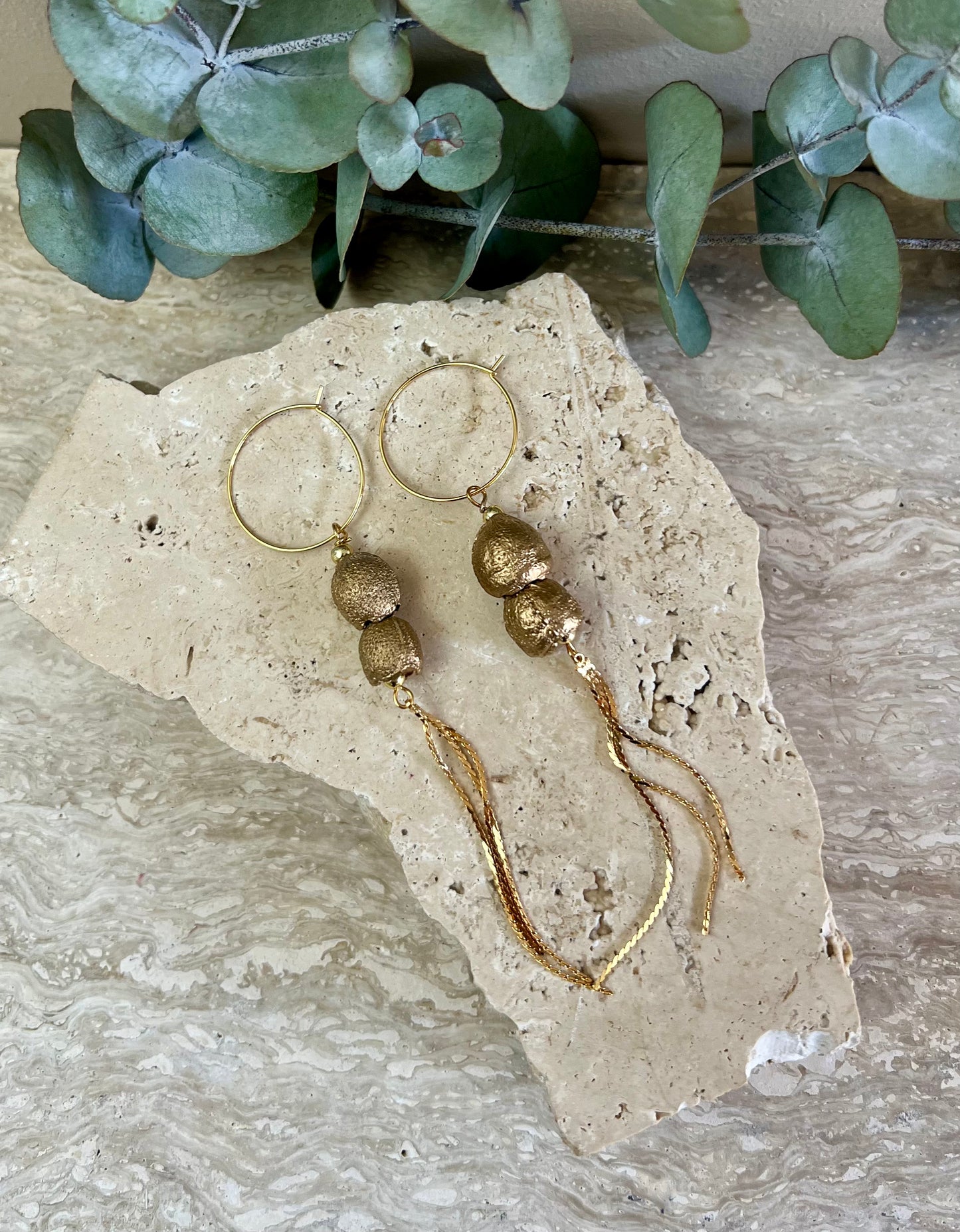 Gold beaded earrings on a textured stone surface with eucalyptus leaves in the background.