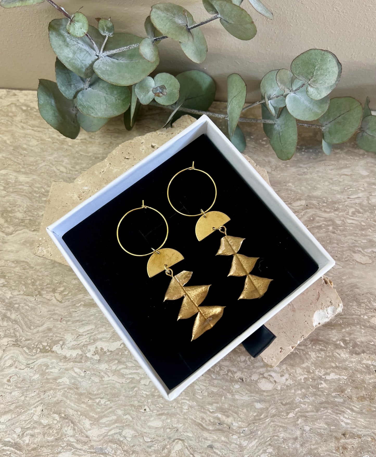 Gold earrings in a box with eucalyptus leaves on a stone surface