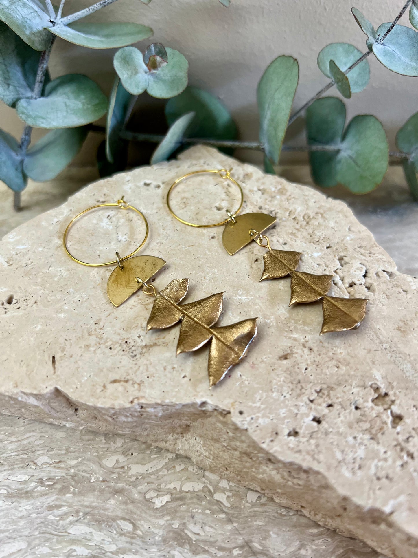 Gold hoop earrings with leaf-like charms on a stone surface with greenery in the background