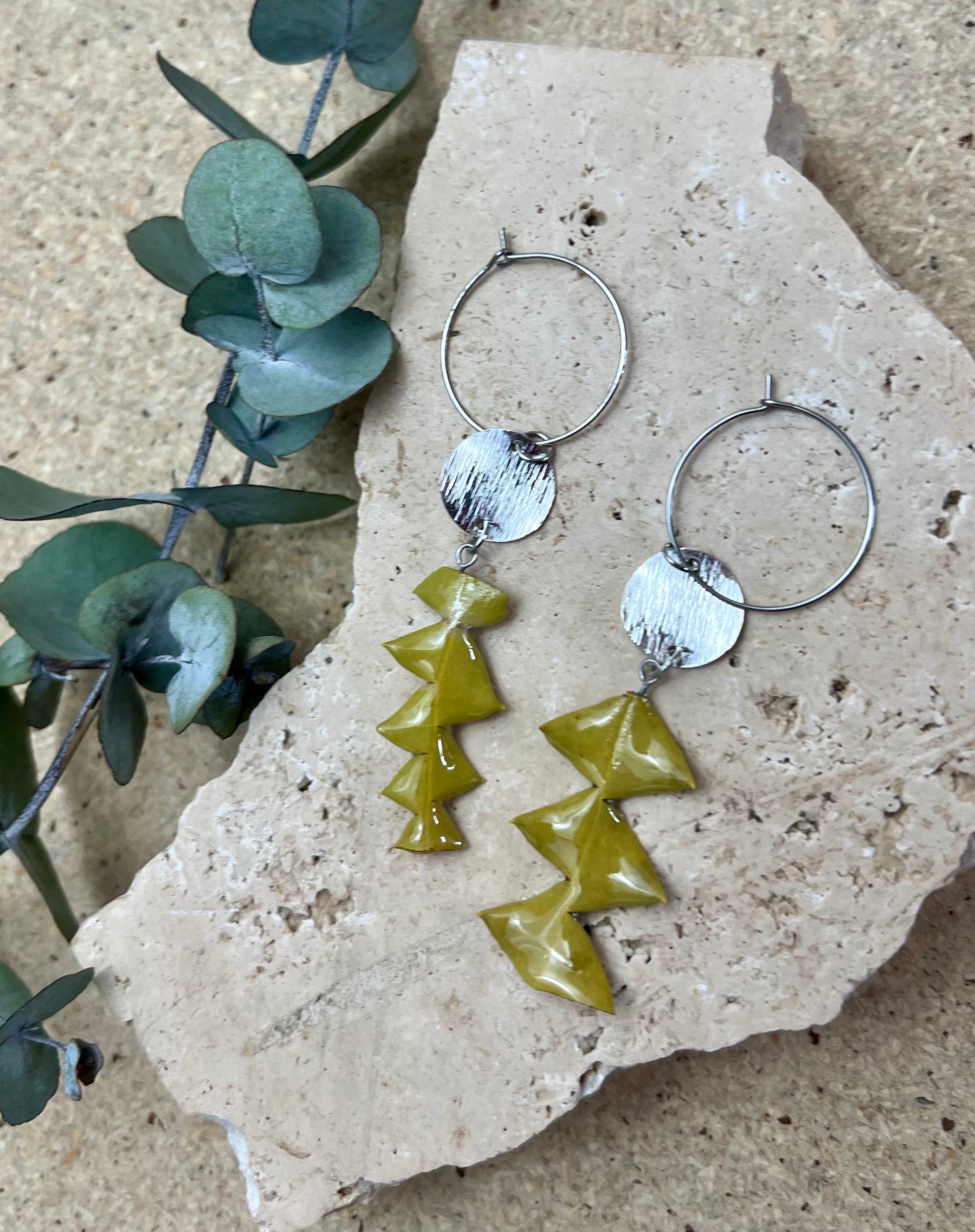 Pair of hoop earrings with green and silver geometric pendants on a stone surface.