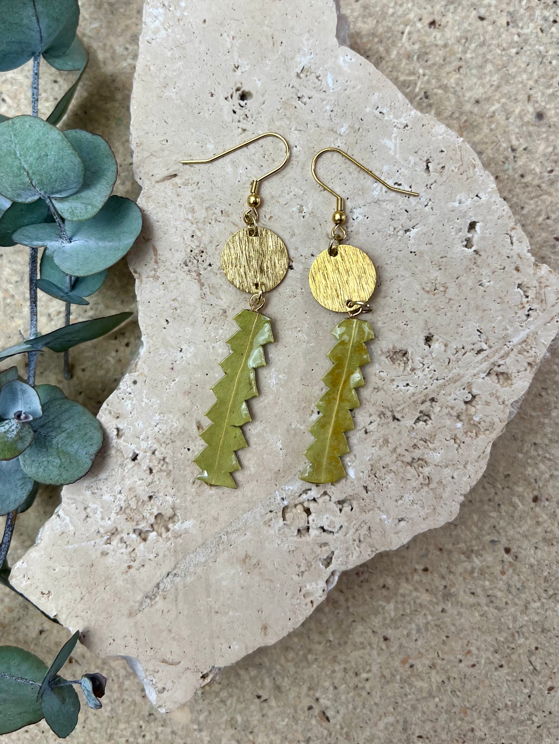 Gold and green leaf-shaped earrings on a stone surface with eucalyptus leaves.