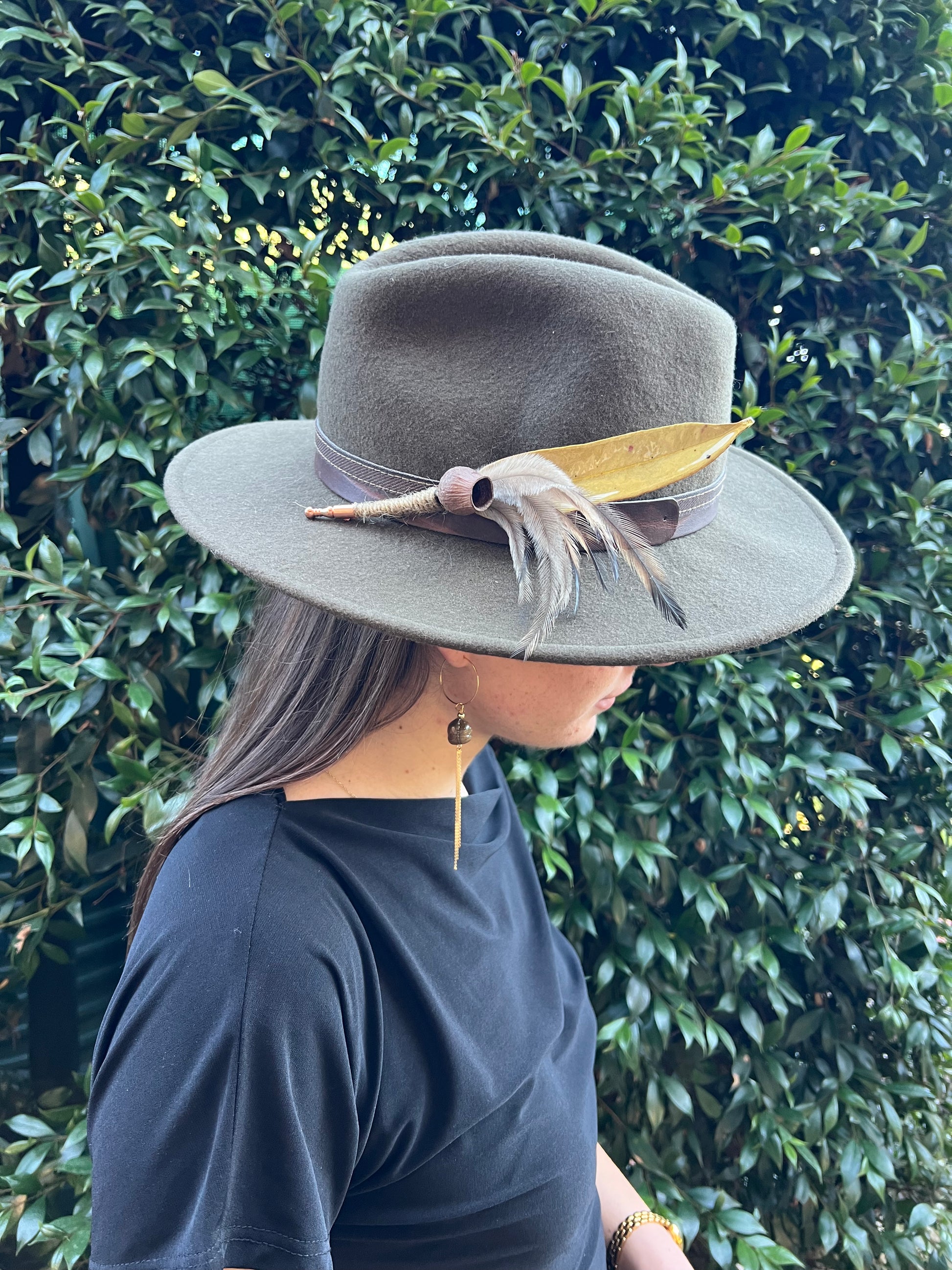 Person wearing a gray hat with a feather against a green hedge background