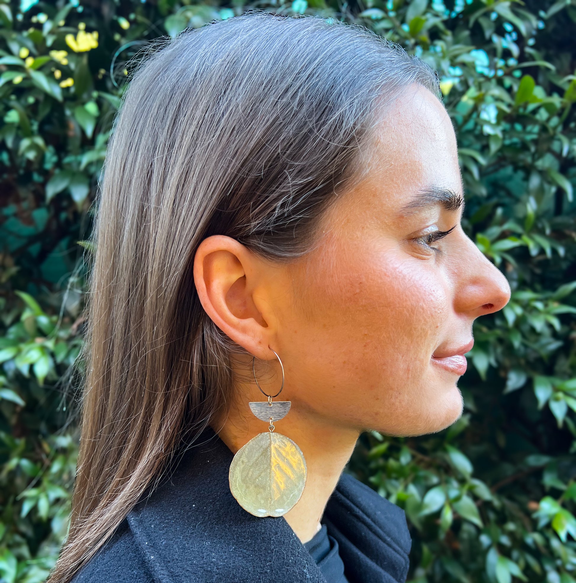 Woman wearing a large, leaf-shaped earring with a green leaf pattern against a blurred natural background.